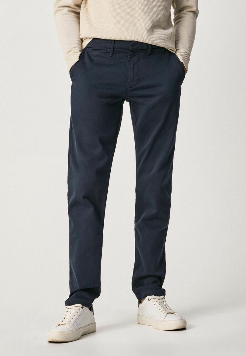 Homme portant un pantalon chino bleu marine foncé, un pull beige et des baskets blanches, debout les mains dans les poches devant un fond uni.