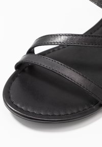 Gros plan d'une zone des orteils d'une sandale en cuir noir avec deux fines sangles cousues se croisant sur la semelle.