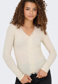 Beige ribbattu pitkähihaisekaulus, jossa on V-pääntie, pitsireunus keskellä ja aaltoileva helma. Teksturoitu kangas, joka istuu tiiviisti.