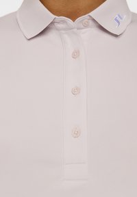 Jasnofioletowa koszulka polo z żebrowanym kołnierzykiem, pięcioma guzikami i małym niebieskim logo na kołnierzyku. Gładka, oddychająca tekstura materiału.