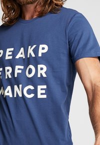 Modrá bavlněná tričko s velkými bílými blokovými písmeny nápisem "SPEAKP PERFORMANCE." Krátké rukávy a kulatý výstřih.