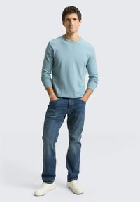 Lichtblauw shirt met lange mouwen gecombineerd met donkerblauwe spijkerbroek en witte sneakers. De stof lijkt zacht; het shirt heeft geribbelde manchetten en een ronde hals.