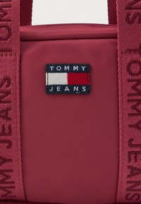Sötétvörös szövet táska hímzett Tommy Jeans logóval és széles, azonos színű pántokkal, amelyeken a "Tommy Jeans" felirat van szőve.