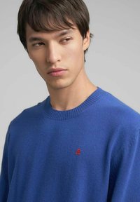 Maglione blu realizzato in materiale morbido, con scollo a girocollo a coste e un piccolo logo rosso ricamato sul petto a sinistra.