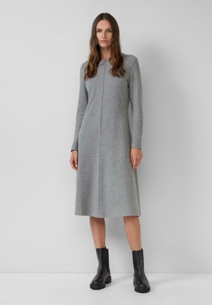 Frau steht vor einer schlichten Wand, trägt ein langärmliges graues Midikleid und schwarze Stiefeletten, mit glatt herunterhängendem braunem Haar.