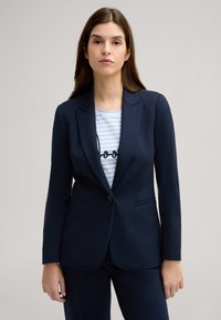 Blazer blu navy con vestibilità su misura, revers a notched e chiusura con un solo bottone. Indossato sopra un top a righe blu chiaro con testo.