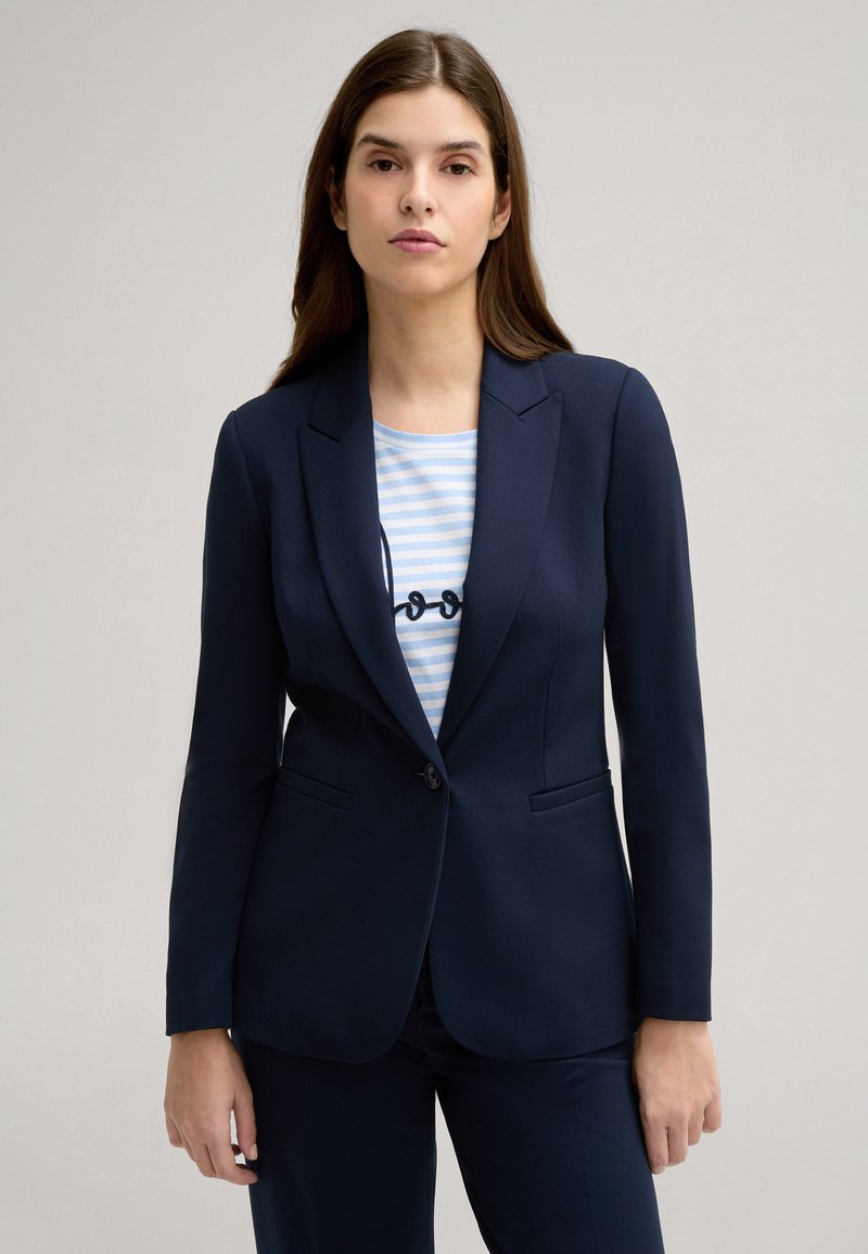 Blazer blu navy con vestibilità su misura, revers a notched e chiusura con un solo bottone. Indossato sopra un top a righe blu chiaro con testo.