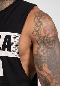 Zwarte mouwloze tanktop met een ruime armsgat. Bevat witte grafische tekst. Geweven tatoeageontwerpen zichtbaar op de bovenarm.