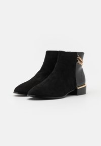 Par de botines de ante y cuero negro con punteras puntiagudas, tacones cuadrados y adornos de cadena dorada y tachuelas en la parte trasera.