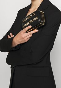 Czarna kopertówka z złotym napisem "LUKSUS TO DYSCYPLINA", charakteryzująca się zorganizowaną konstrukcją i złotymi akcentami sprzętu.