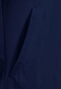 Tessuto blu navy con una superficie liscia, dotato di una tasca laterale con dettagli di cucitura discreti. Il design è minimalista e utilitario.