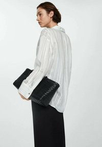 Witte gestreepte blouse met lange mouwen, zwarte gevlochten clutch tas en lange zwarte rok, gezien van achteren.