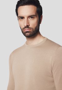 Beżowy sweter dzianinowy z okrągłym dekoltem. Żebrowane mankiety i dół podkreślają gładką strukturę. Minimalistyczny design, brak widocznych wzorów lub logo.