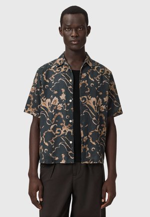 Junger Mann, der ein kurzärmeliges Hemd mit braunen Blumenmustern über einem schwarzen T-Shirt und dunkler Hose vor einem einfarbigen Hintergrund trägt.