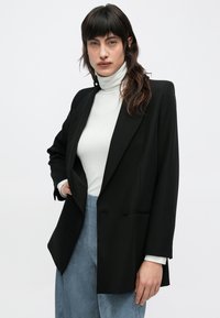 Sort skræddersyet blazer med dobbeltradet lukning, hakkede revers og tekstureret stof, båret over en hvid rullekrave og blå kordbuks.