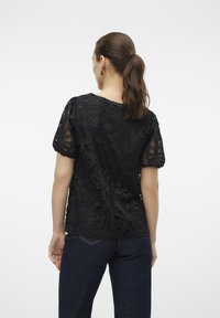 Sort blusel med blonder, der har et blomstermotiv med udskæring, korte puffærmer og en rund halsudskæring, parret med mørke denimjeans.