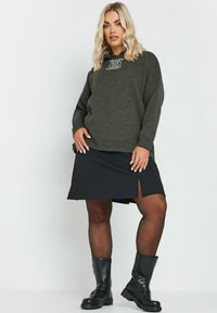 Grön tröja med broderad logotyp på bröstet, kombinerad med en svart kjol med en slits på sidan. Svarta knähöga stövlar fulländar outfiten.