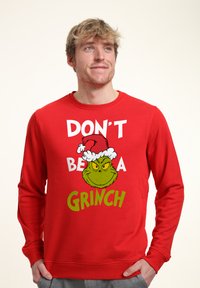 Felpa rossa con grafica del Grinch in un cappello da Babbo Natale e testo "NON ESSERE UN GRINCH" in bianco e verde. Tessuto morbido, maniche lunghe.