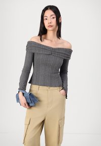 Off-the-shoulder gray ribbed top with pleated accents, long sleeves, and fitted design, paired with beige cargo pants and a denim accessory.