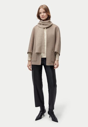 Frau steht, trägt beigen Schal und Jacke, cremefarbene Bluse, schwarze Lederhose und schwarze spitze Stiefel mit hohen Absätzen vor einfachem Hintergrund.