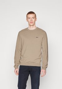 Tan knitted sweater with a round neck and ribbed cuffs. Features a small black logo on the chest, paired with dark blue pants.
