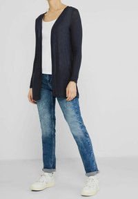 Osoba oblečená v námořnickém otevřeném kardiganu, bílém topu, zahnutejch modrých džínách a bílých teniskách na světlém jednobarevném pozadí.