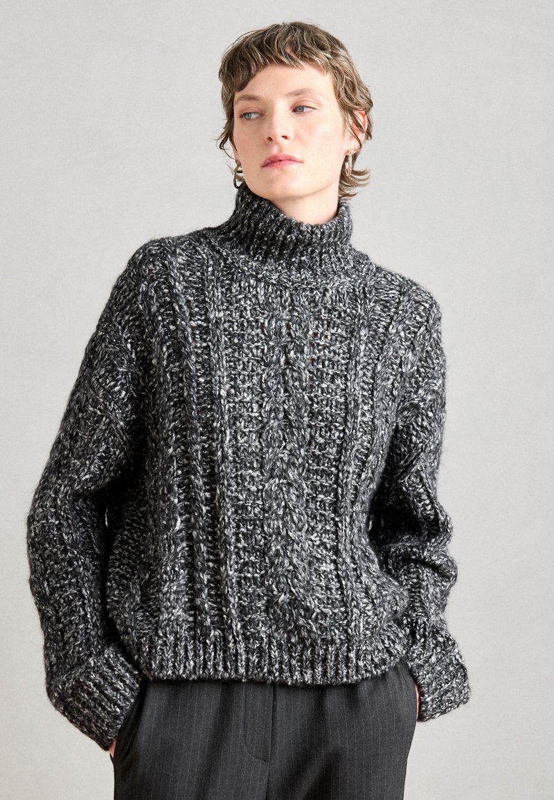 Pullover a collo alto in maglia grossa di colore grigio scuro con motivo testurizzato, vestibilità ampia e polsini e fondo a coste. Indossato sopra pantaloni sartoriali.