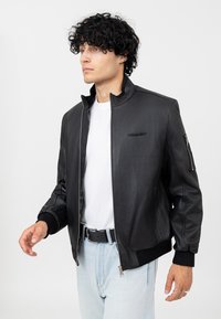 Giubbotto bomber in pelle nera con colletto alto, chiusura con zip e polsi a costine. Presenta un piccolo dettaglio del logo sul petto e una tasca laterale.