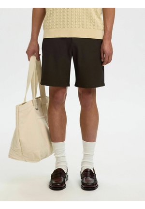 Personne portant un short noir, des chaussettes blanches et des mocassins marron foncé, tenant un grand sac fourre-tout beige dans la main gauche.