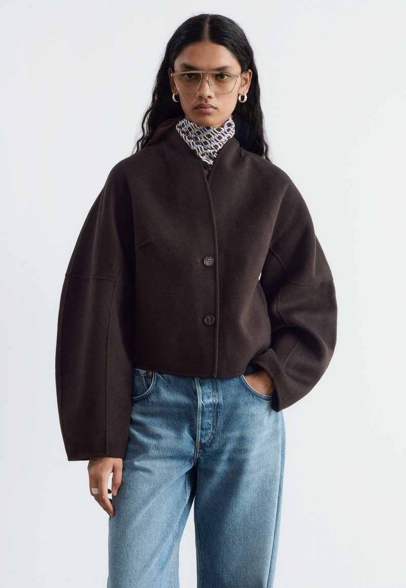 Femme aux longs cheveux foncés portant une veste marron foncé oversize, une écharpe à motifs, des lunettes transparentes, des boucles d'oreilles créoles et un jean bleu clair, mains dans les poches.