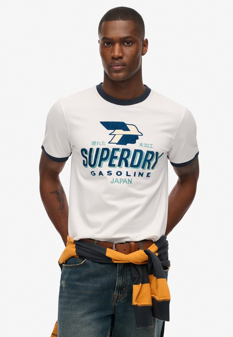 Hvid kortærmet t-shirt med marineblå krave, med "SUPERDRY" grafik i blå og grøn. Båret med slidte denimjeans og en stribet sweater bundet om taljen.