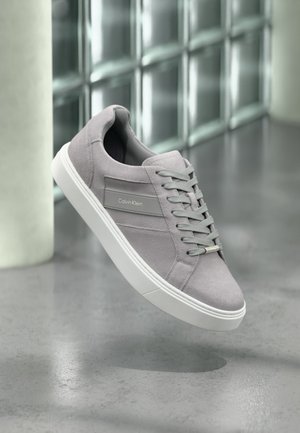 Baskets en suède gris de Calvin Klein avec semelle et lacets blancs, flottant au-dessus d'un sol en béton poli près de blocs de verre.