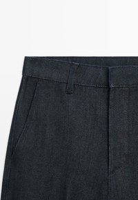 Dark denim trousers feature a straight-cut design, a fitted waistband, side pockets, and subtle stitching along the edges.