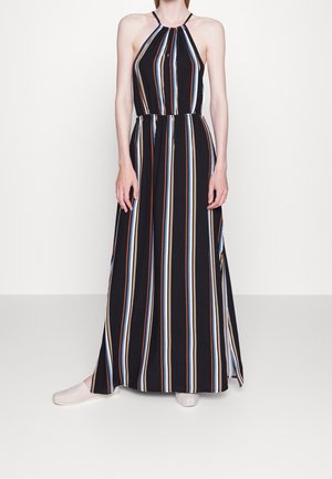 Vrouw draagt een mouwloze zwarte maxi-jurk met verticale blauwe, witte en bruine strepen, gecombineerd met lichtgekleurde instapschoenen.