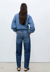 Niebieska kurtka pikowana z bufiastymi rękawami, w połączeniu z niebieskimi dżinsami o wysokim stanie i czarnymi sandałami, widok z tyłu.