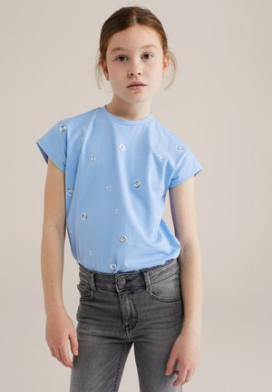 Maglietta in cotone blu chiaro con maniche corte, caratterizzata da dettagli a occhio circolare argentati sparsi sulla parte anteriore. Abbinata a jeans grigi.