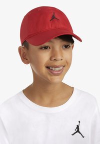 Cappellino da baseball rosso in tessuto, con il logo Jumpman nero ricamato sul davanti. Indossato con una maglietta bianca.
