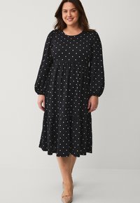 Vestido negro de lunares con mangas largas, escote redondo, cuerpo fruncido, falda en capas y textura de tela suave. Diseño de longitud media.