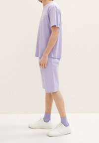 Conjunto de camiseta y pantalones cortos de color lavanda, hecho de tejido ligero. La camiseta tiene un cuello redondo y un corte relajado; los pantalones cortos son de longitud hasta la rodilla. Zapatillas blancas y calcetines morados.