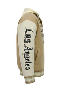 Beige en crèmekleurige varsityjas met geribbelde manchetten, met "Los Angeles" in grote zwarte letters op de mouw en een ritssluiting.