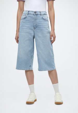 Φαρδιά τζιν σορτς σε ανοιχτό μπλε χρώμα με ψηλή μέση, συνδυασμένα με λευκές κάλτσες και λευκά αθλητικά παπούτσια, φορεμένα με λευκό κοντομάνικο τοπ.