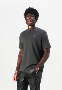 T-shirt gris foncé à manches courtes en coton doux, avec un petit logo sur la poitrine. Porté avec un pantalon cargo en cuir noir avec des poches.