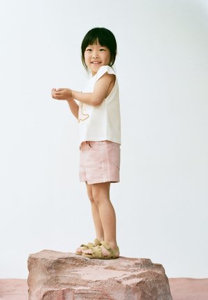 Jeune fille debout sur un rocher, portant un haut blanc sans manches, un short rose et des sandales vertes, souriante et tenant ses mains ensemble.