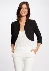 Schwarze cropped Jacke mit 3/4-Ärmeln, tailliertem Design und glatter Textur, getragen über einem weißen Oberteil mit Spitzenakzenten und Knöpfen.