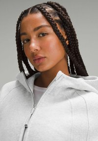 Grå zip-huvtröja med mjuk textur, huva och kontrasterande sömmar. Modellen har flätat hår och bär en vit skjorta under.