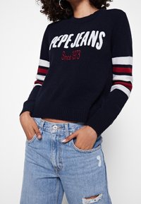 Pull en maille bleu marine avec l'inscription "Pepe Jeans" en texte blanc et rouge, avec des manches rayées multicolores. Porté avec un jean délavé bleu clair.