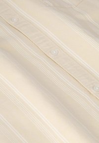 Beige Hemd mit weißem Streifenmuster, mit einem Button-Down-Kragen und weißen Knöpfen entlang der Leiste; glattes Textil. 