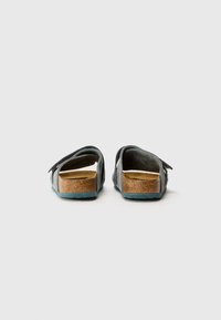 Sandales en suède gris avec semelle en liège, sangle ajustable et semelle en caoutchouc teal. Design ouvert à l'arrière, révélant la texture naturelle du matériau.