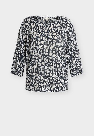 Blouse à motif floral noir et blanc avec manches longues et encolure ronde, présentée sur un fond blanc uni.
