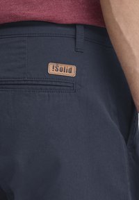 Navy blue shorts with a smooth texture, featuring a leather patch with "iSolid" branding and a back pocket detailing.